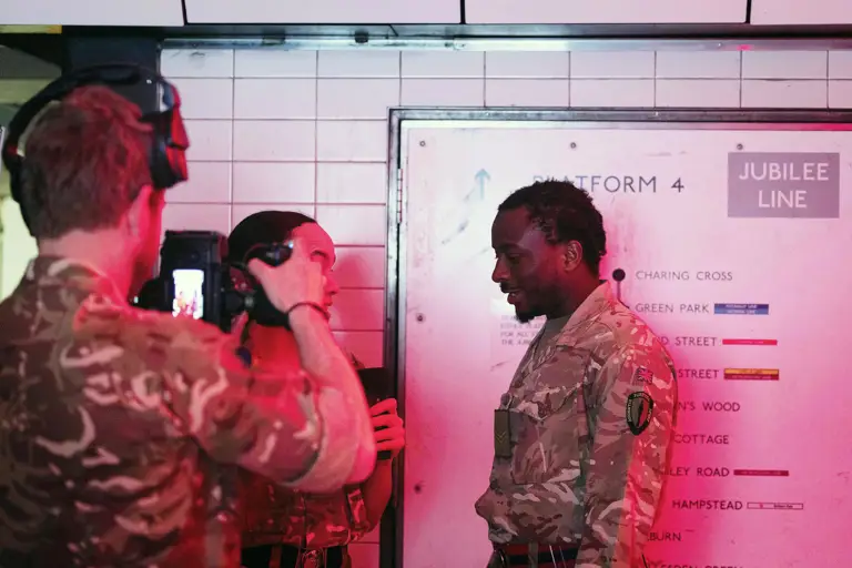 A group of individuals in camouflage uniforms stand on a subway platform beneath a