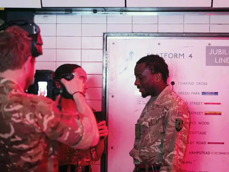 A group of individuals in camouflage uniforms stand on a subway platform beneath a