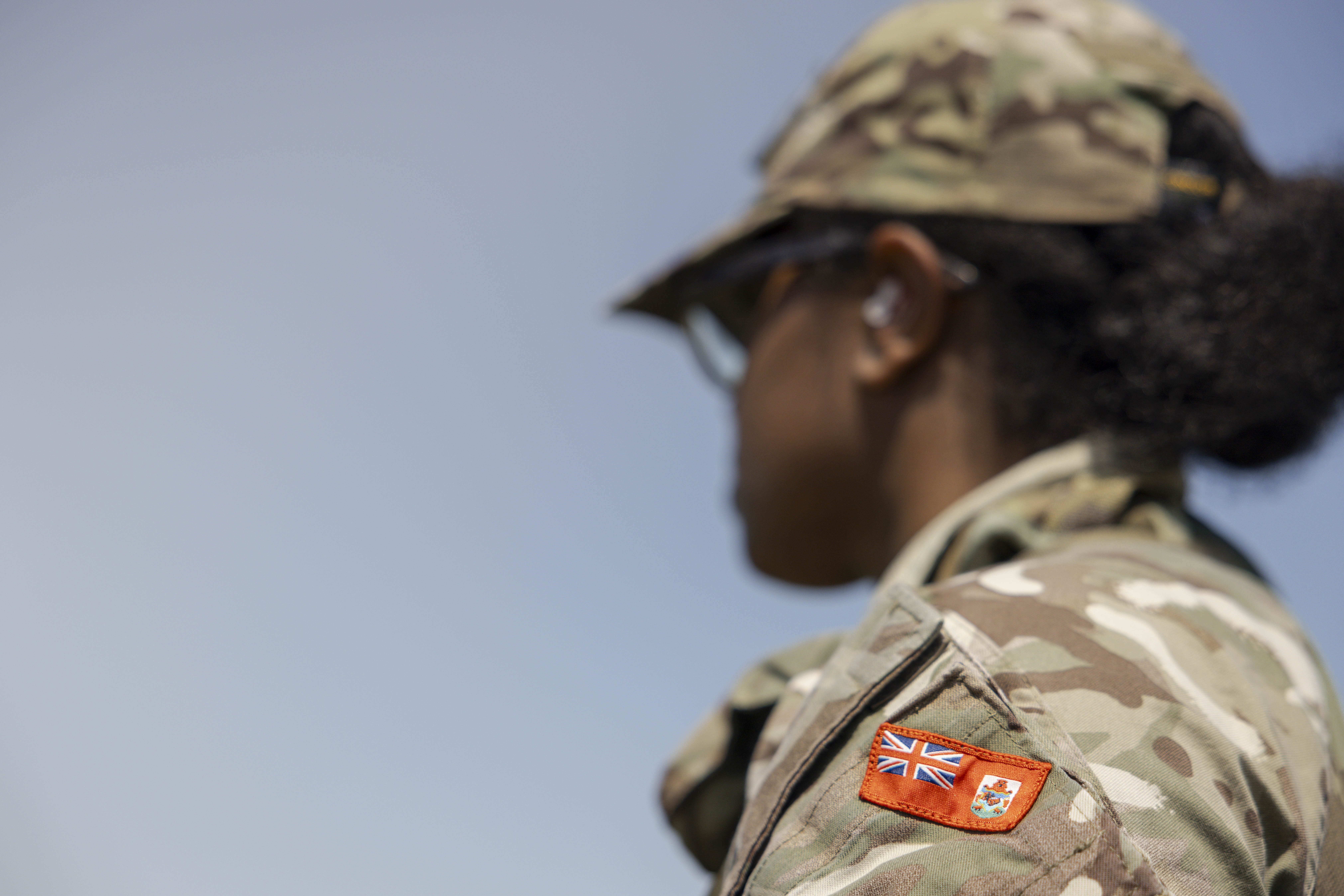 An individual in camouflage military attire stands outdoors. The uniform sleeve features a patch.