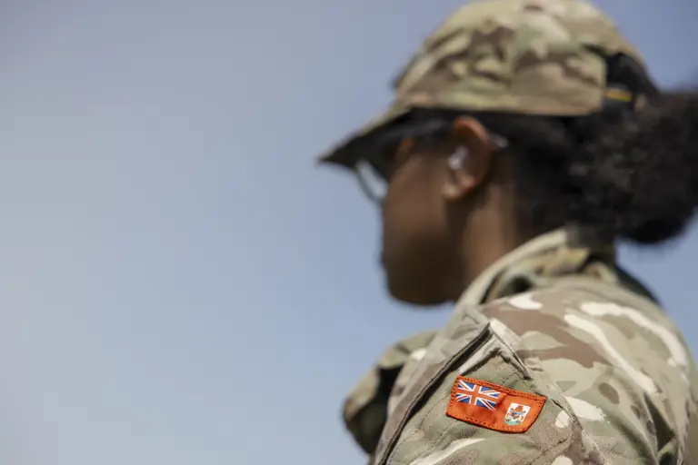 An individual in camouflage military attire stands outdoors. The uniform sleeve features a patch.