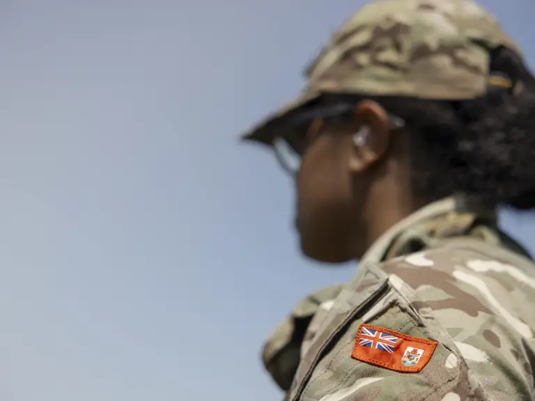 An individual in camouflage military attire stands outdoors. The uniform sleeve features a patch.