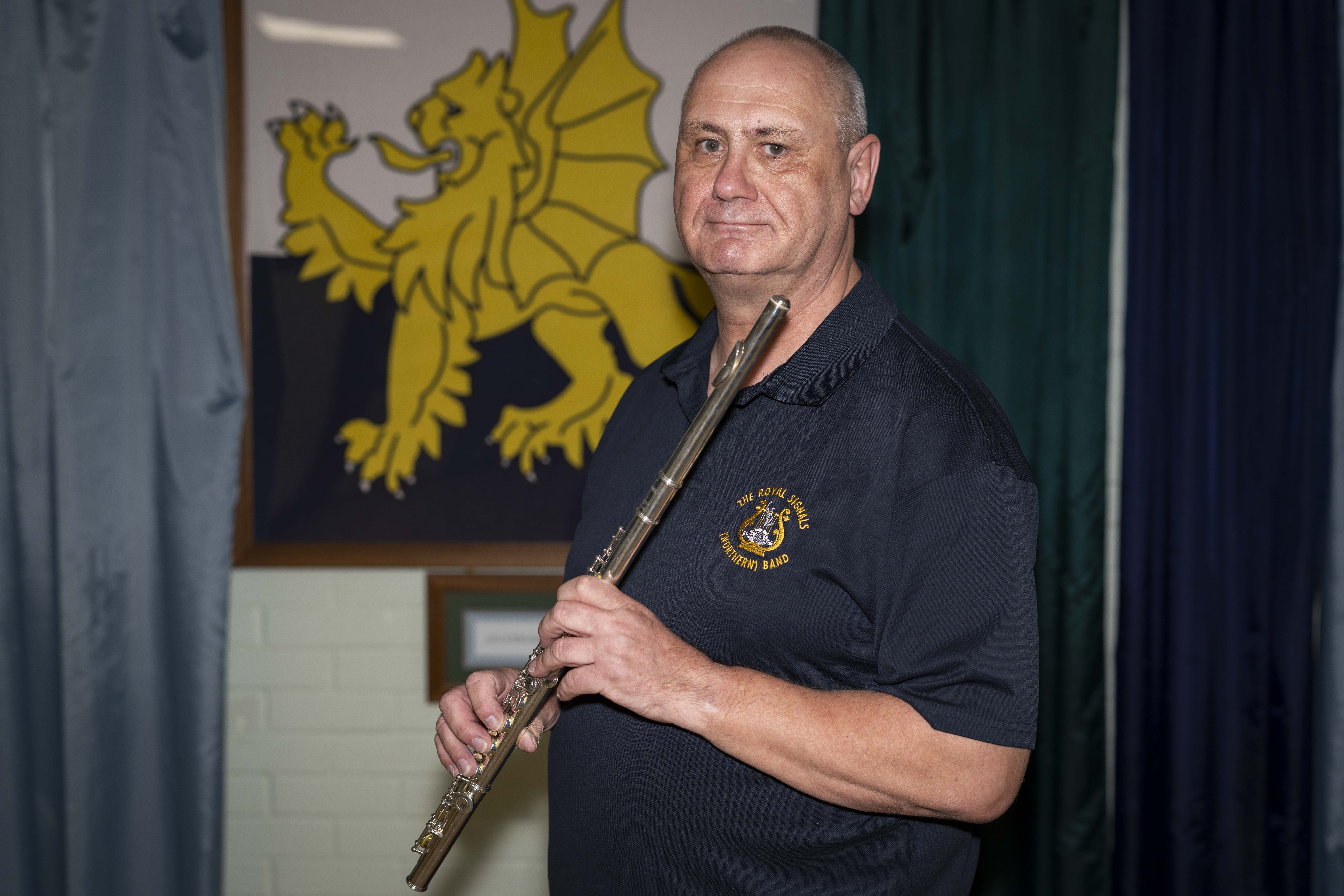A person holding a flute stands in front of a framed emblem featuring a yellow dragon on a dark background. 