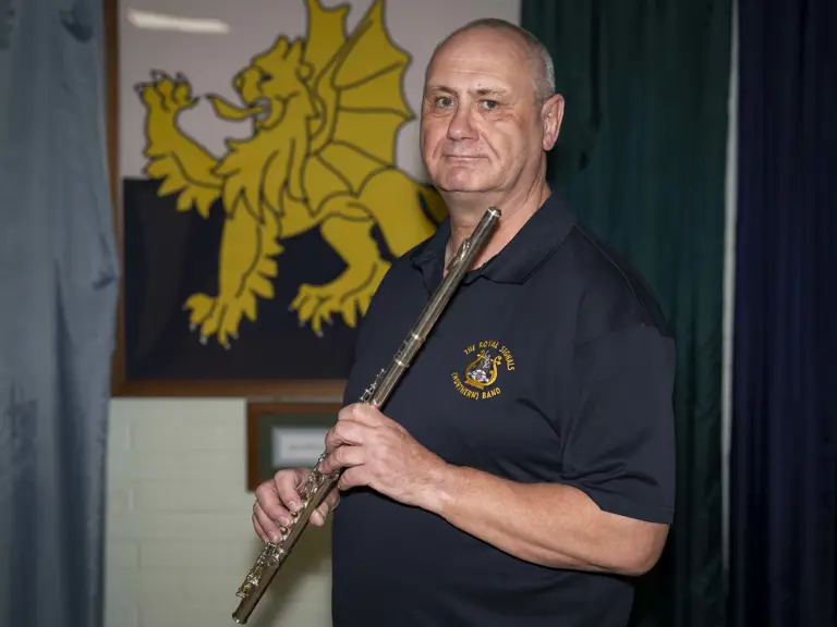 A person holding a flute stands in front of a framed emblem featuring a yellow dragon on a dark background.