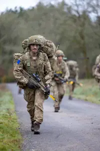 A group of soldiers in camouflage uniforms and heavy backpacks walk along a rural road. Each has a rifle.