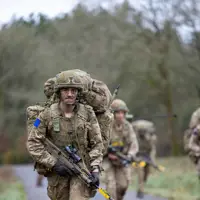 A group of soldiers in camouflage uniforms and heavy backpacks walk along a rural road. Each has a rifle.