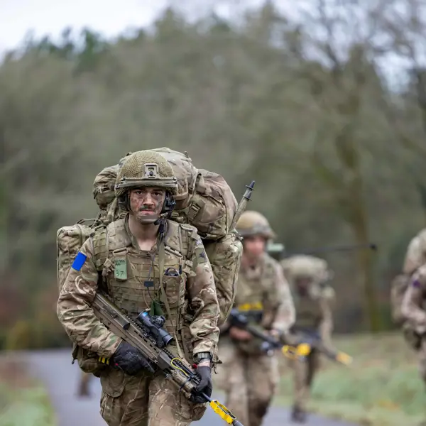 A group of soldiers in camouflage uniforms and heavy backpacks walk along a rural road. Each has a rifle.
