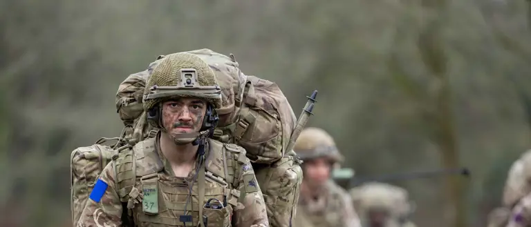 A group of soldiers in camouflage uniforms and heavy backpacks walk along a rural road. Each has a rifle.