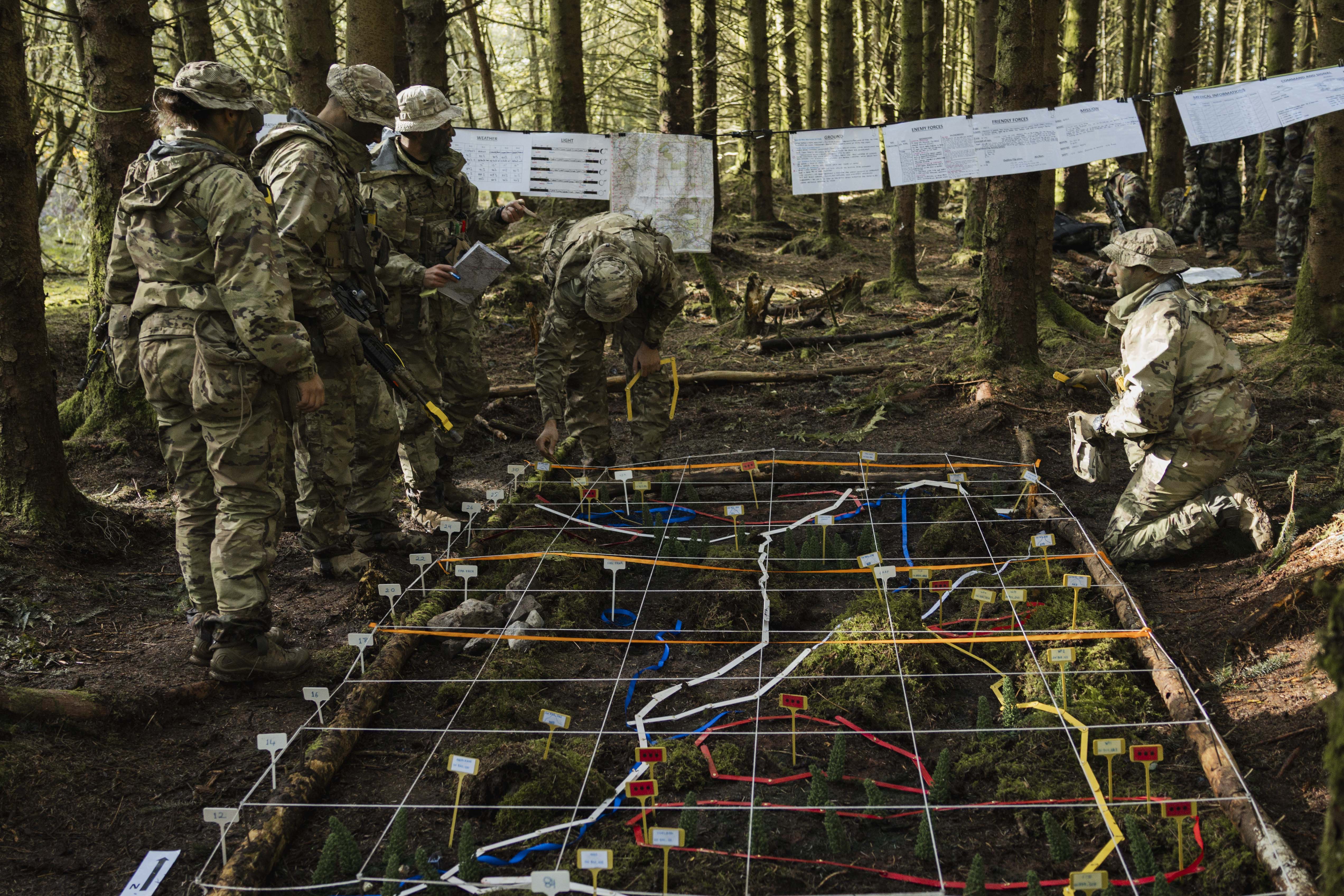 Soldiers in camouflage inspect a terrain model in a forest, featuring grid lines and colourful markers. Maps hang from trees, creating a focused atmosphere.