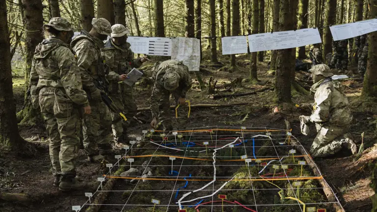 Soldiers in camouflage inspect a terrain model in a forest, featuring grid lines and colourful markers. Maps hang from trees, creating a focused atmosphere.