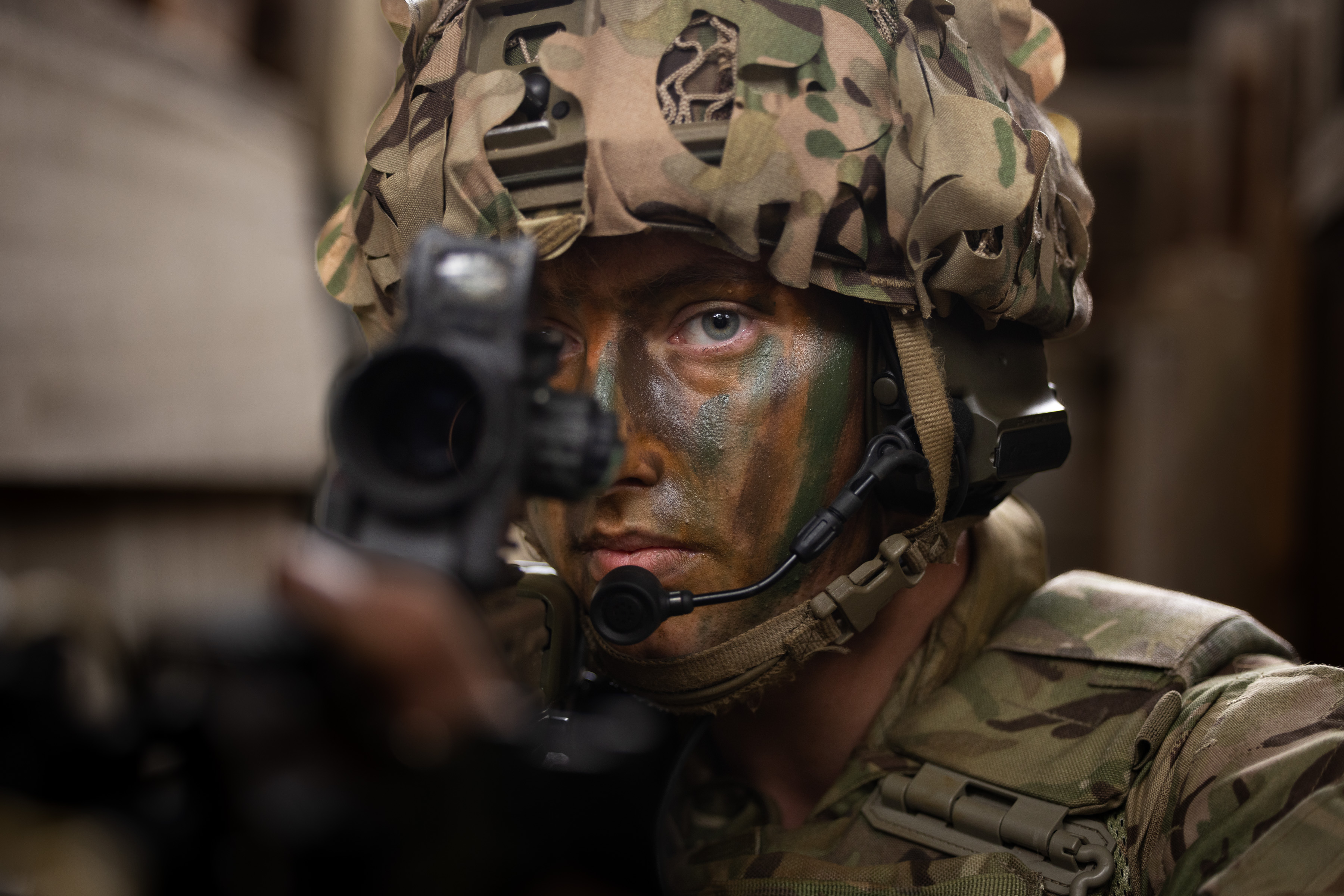 Close up of a soldier's face, wearing camouflage cream, a helmet and looking down the site of his rifle.
