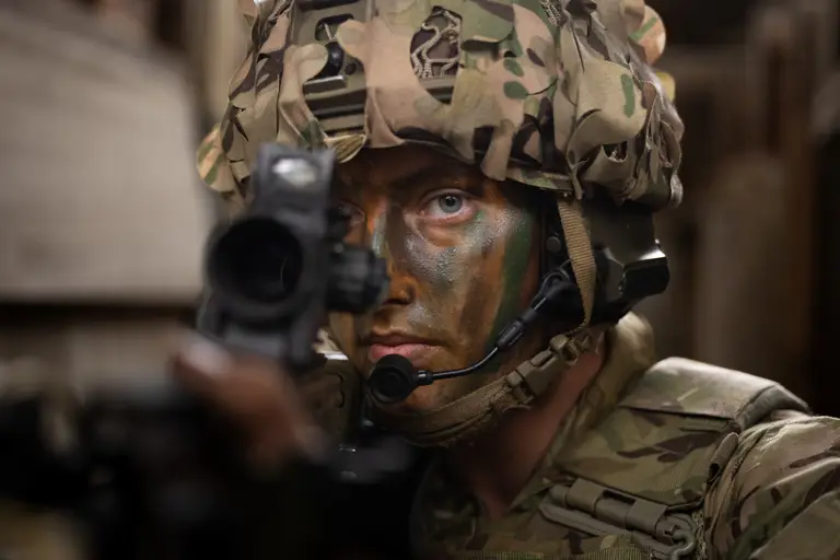 Close up of a soldier's face, wearing camouflage cream, a helmet and looking down the site of his rifle.