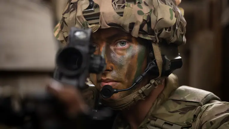 Close up of a soldier's face, wearing camouflage cream, a helmet and looking down the site of his rifle.