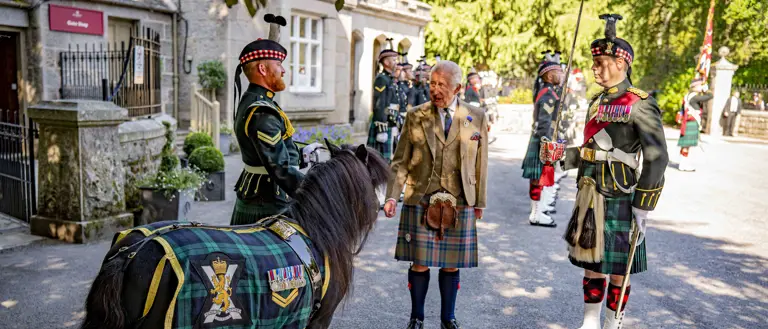 Scottish soldiers in traditional military dress stand with His Majesty The King in a kilt beside a pony draped in tartan.