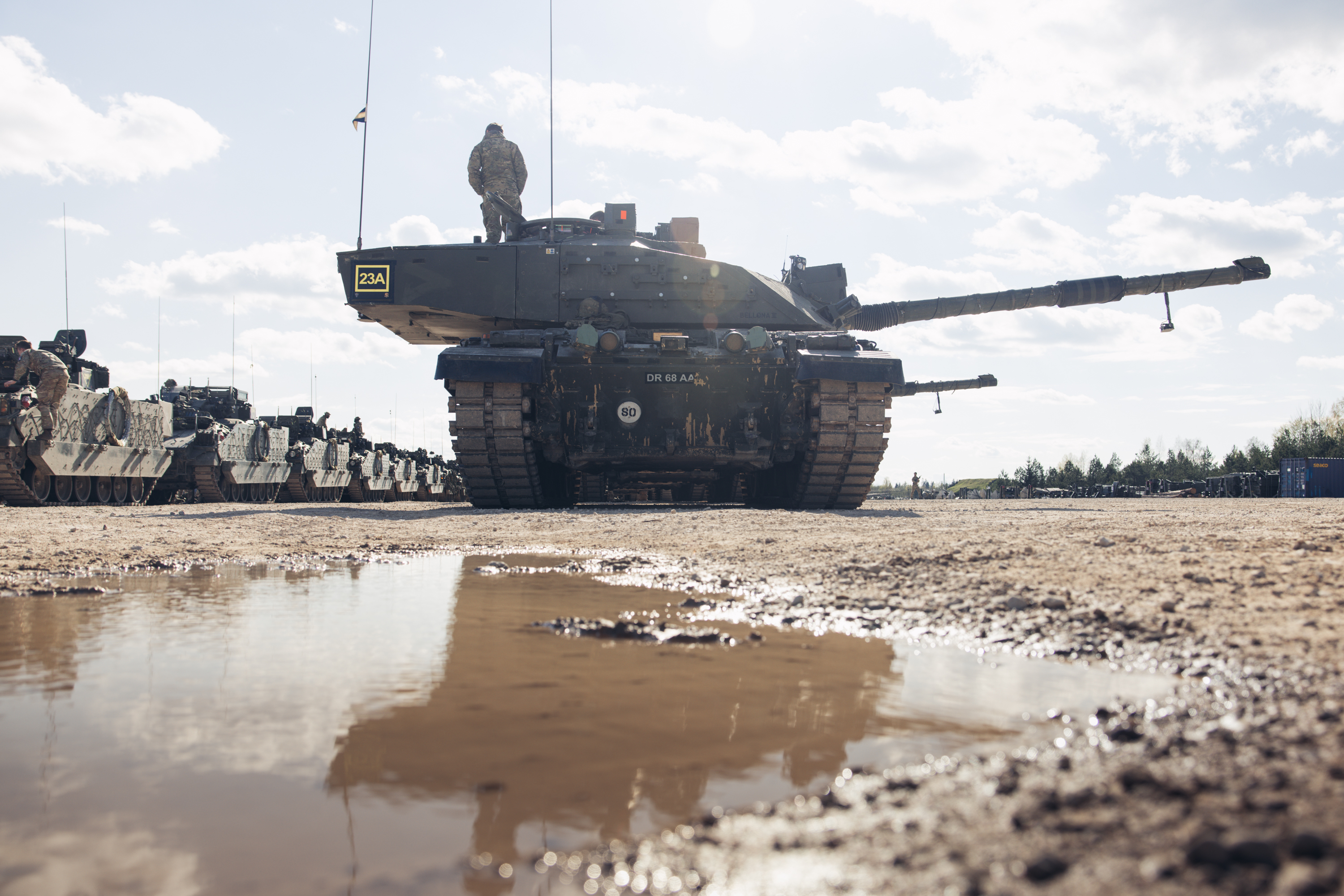 Military tank parked on a muddy ground with a soldier standing on top under a partly cloudy sky.