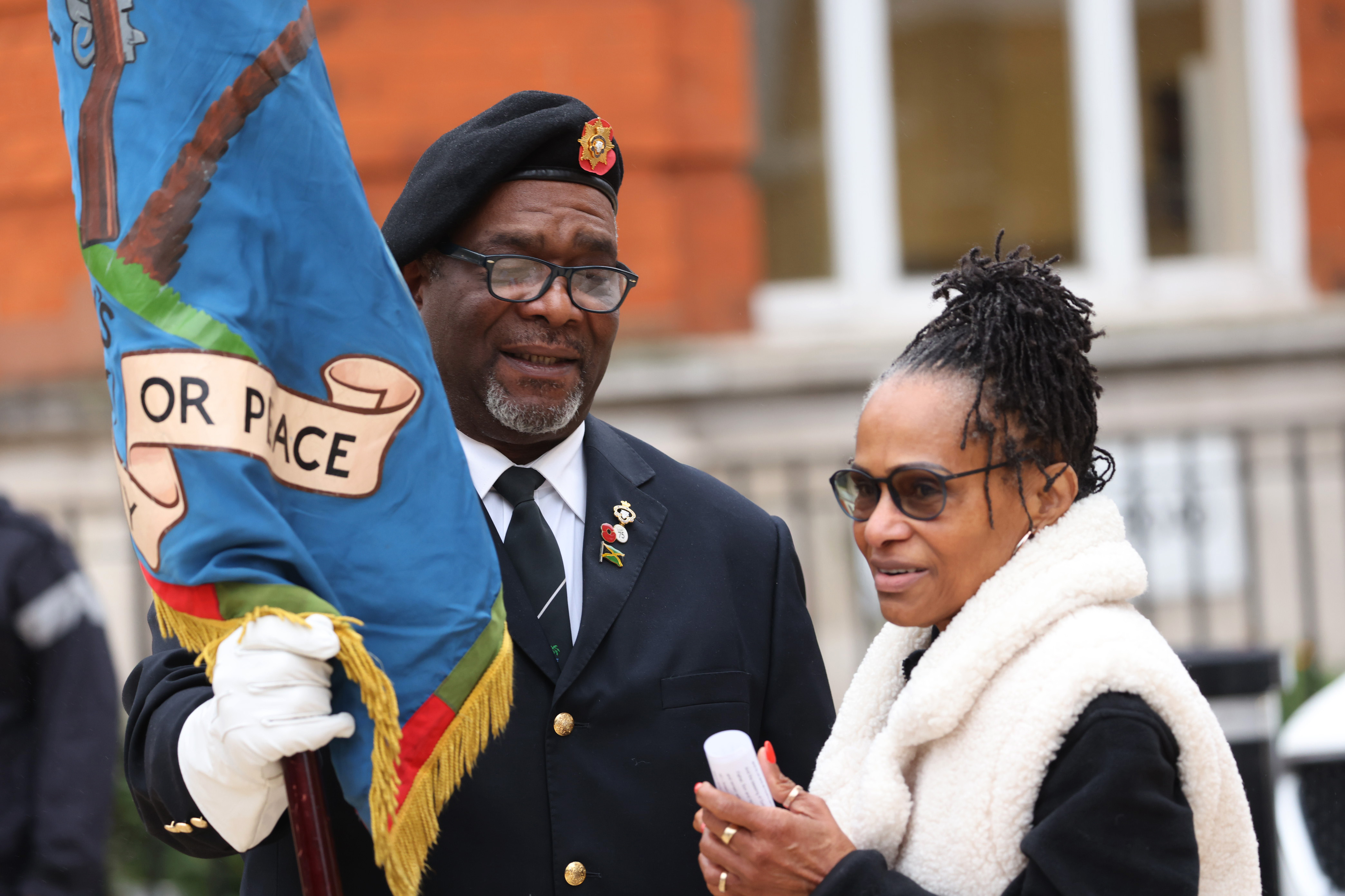 Two people stand, with the person on the left wearing a dark suit and holding a flag.