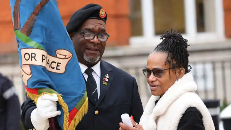 Two people stand, with the person on the left wearing a dark suit and holding a flag.