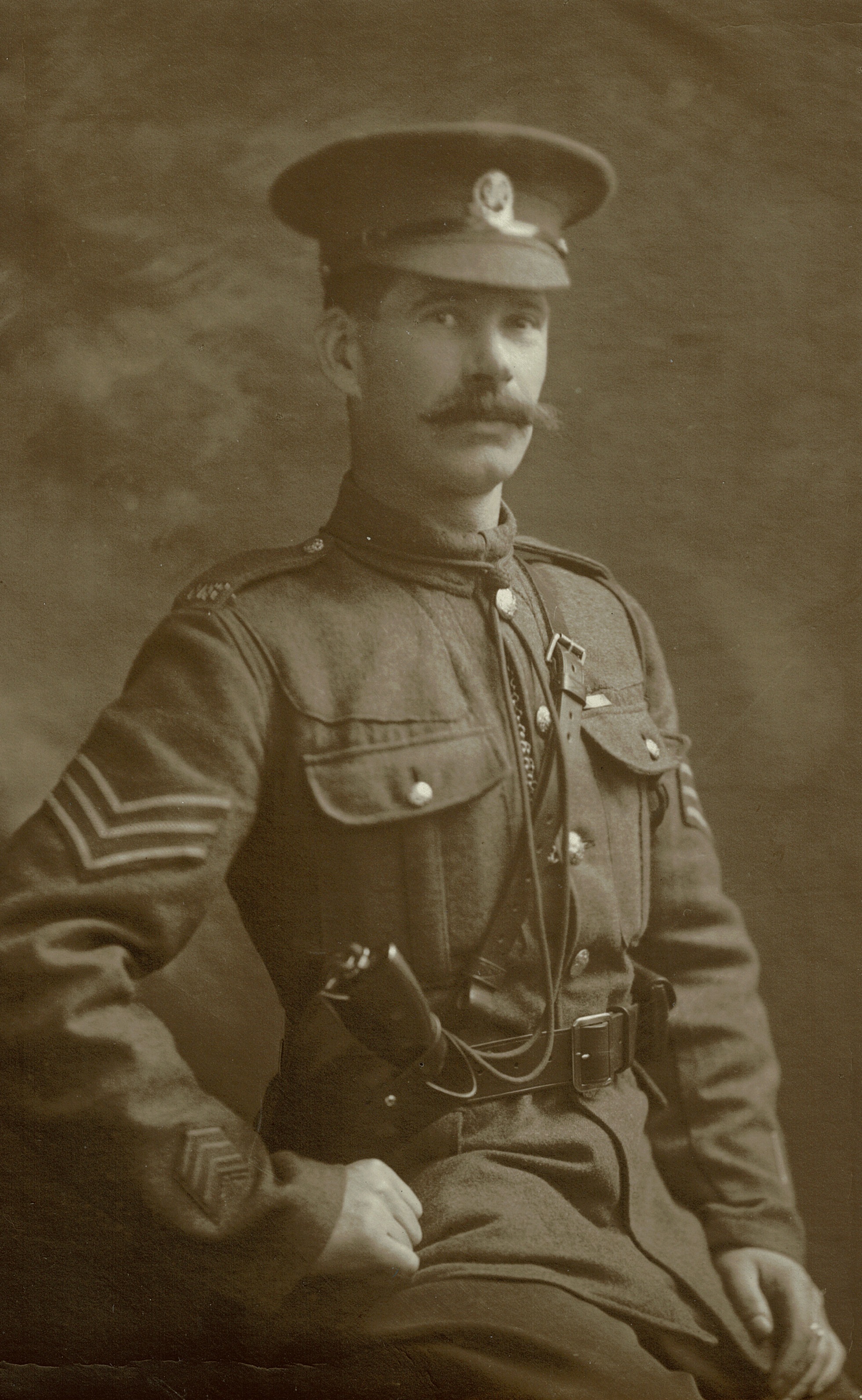 A sepia photo of a soldier from the early 20th century. He wears a uniform with sergeant stripes, a peaked cap, and has a confident expression.