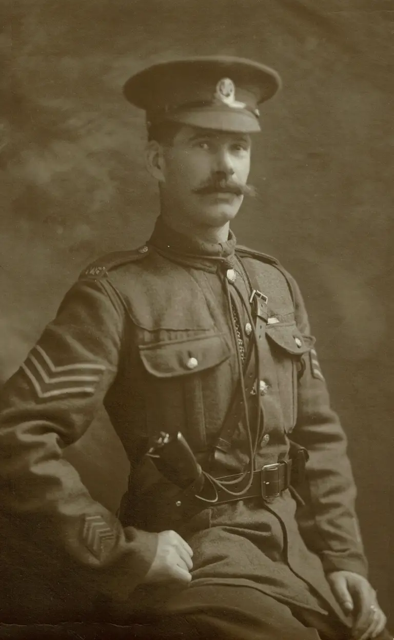 A sepia photo of a soldier from the early 20th century. He wears a uniform with sergeant stripes, a peaked cap, and has a confident expression.