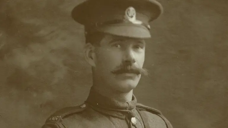 A sepia photo of a soldier from the early 20th century. He wears a uniform with sergeant stripes, a peaked cap, and has a confident expression.