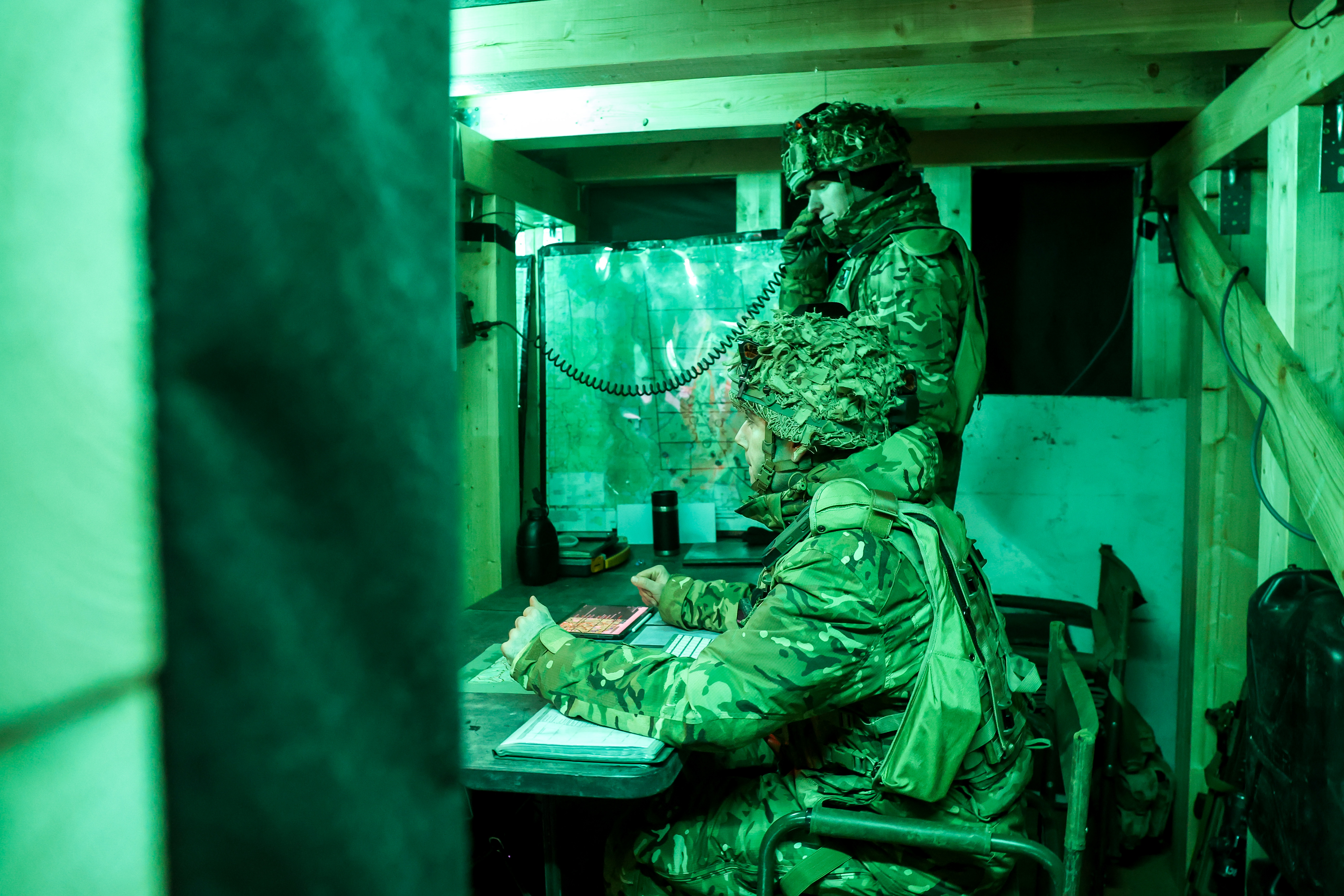 Two soldiers in camouflage gear communicate inside a dimly lit wooden bunker using a field telephone.
