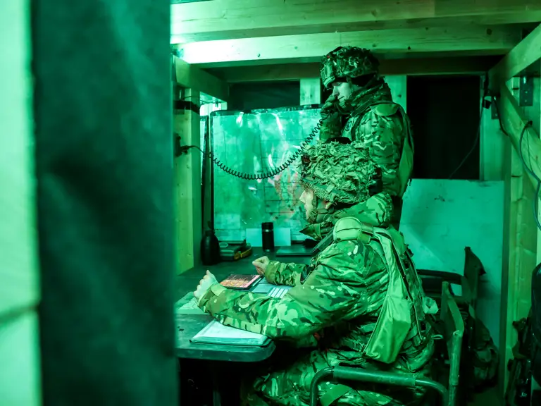 Two soldiers in camouflage gear communicate inside a dimly lit wooden bunker using a field telephone.