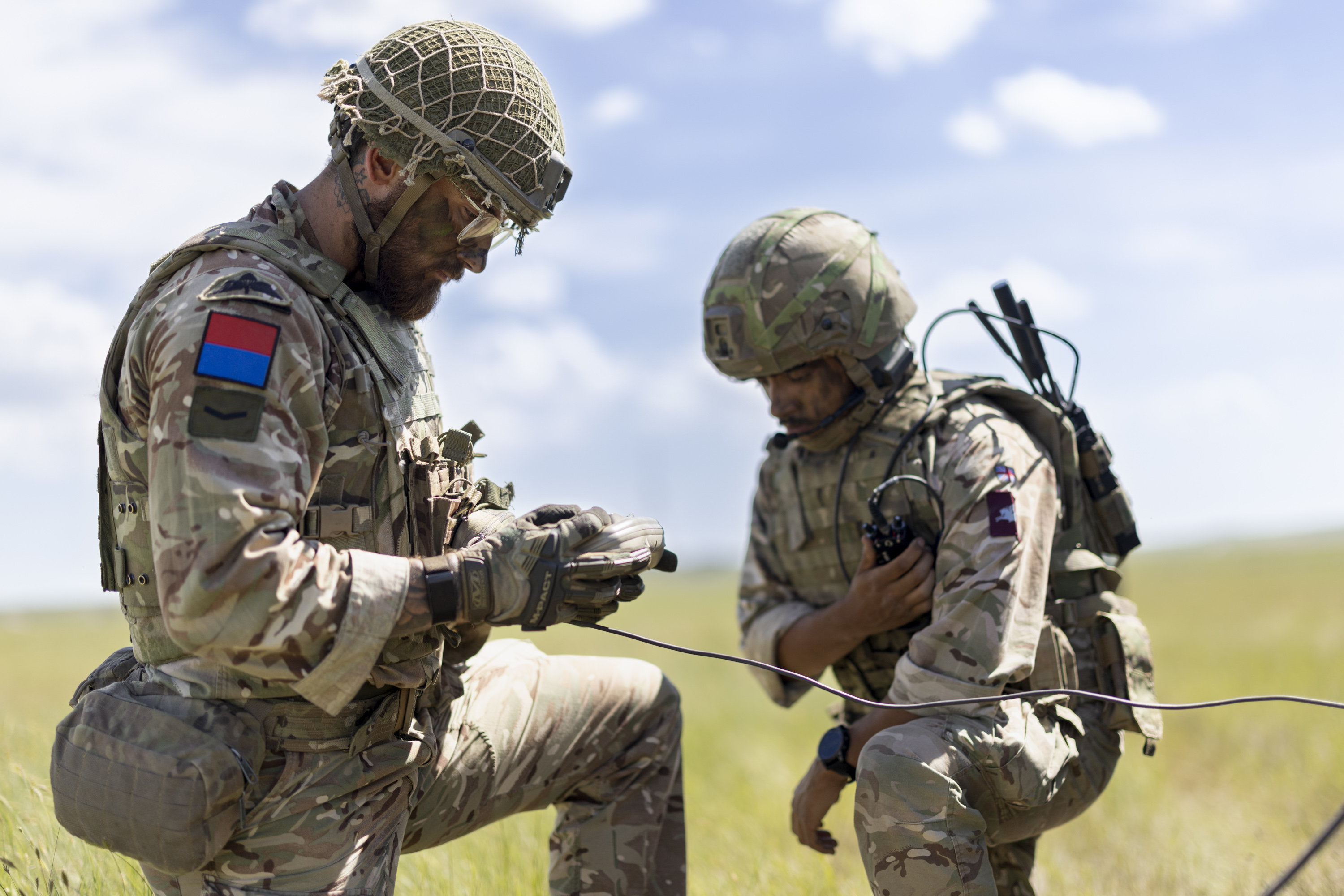 Two soldiers in camouflage uniforms and helmets are working with communication equipment in an open grassy field. One is holding a device, and the other is adjusting gear attached to their attire. The sky is clear with a few clouds.