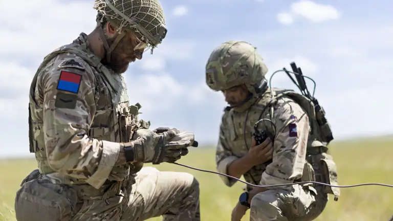 Two soldiers in camouflage uniforms and helmets are working with communication equipment in an open grassy field. One is holding a device, and the other is adjusting gear attached to their attire. The sky is clear with a few clouds.