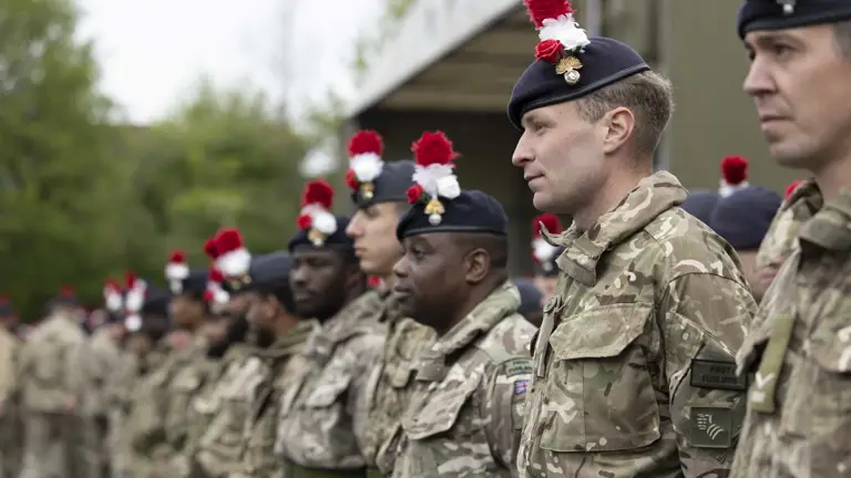 Soldiers in green camouflage uniforms stand on parade. On their headdress they wear a red and white hackle, a red rose and a white rose.