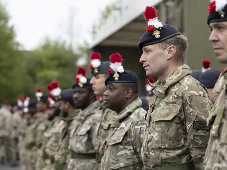 Soldiers in green camouflage uniforms stand on parade. On their headdress they wear a red and white hackle, a red rose and a white rose.