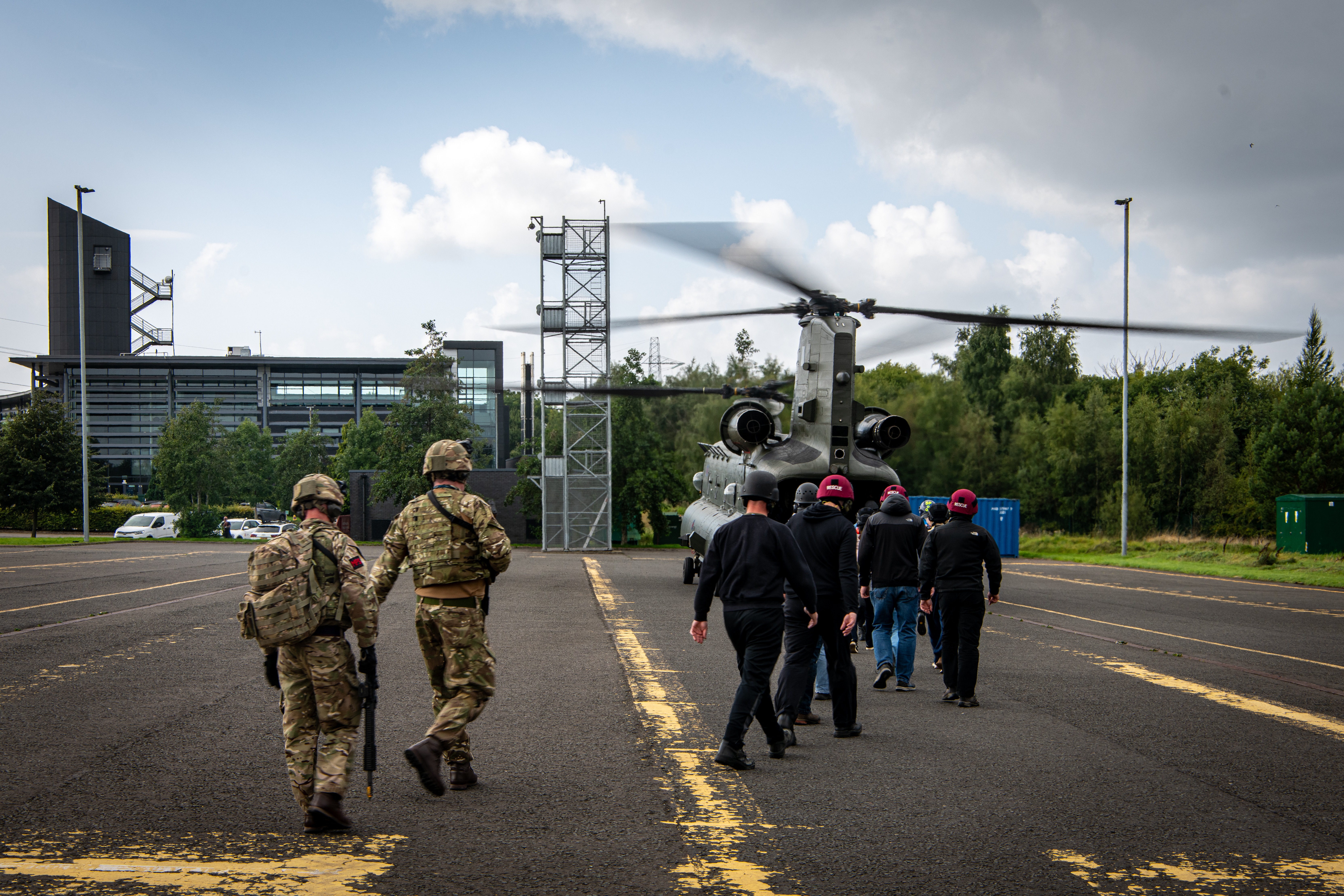 A group of people, including individuals in military uniforms, are walking towards a waiting Boeing CH-47 Chinook helicopter on a tarmac. The surroundings include a modern building and trees under a partly cloudy sky.
