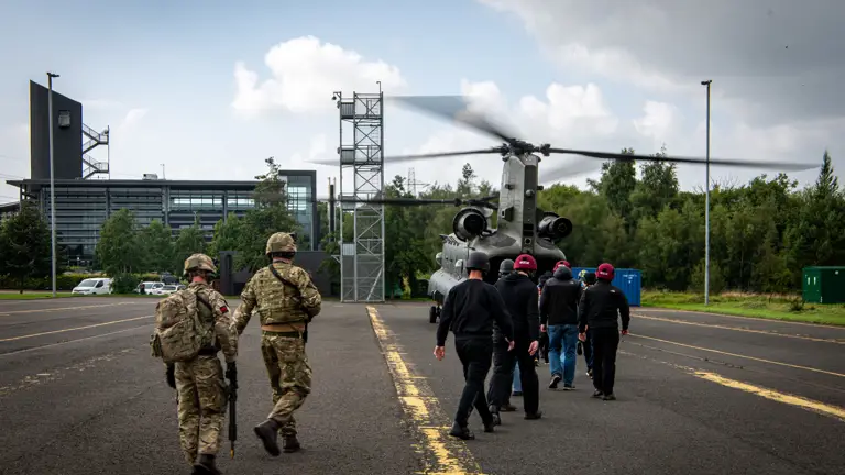 A group of people, including individuals in military uniforms, are walking towards a waiting Boeing CH-47 Chinook helicopter on a tarmac. The surroundings include a modern building and trees under a partly cloudy sky.