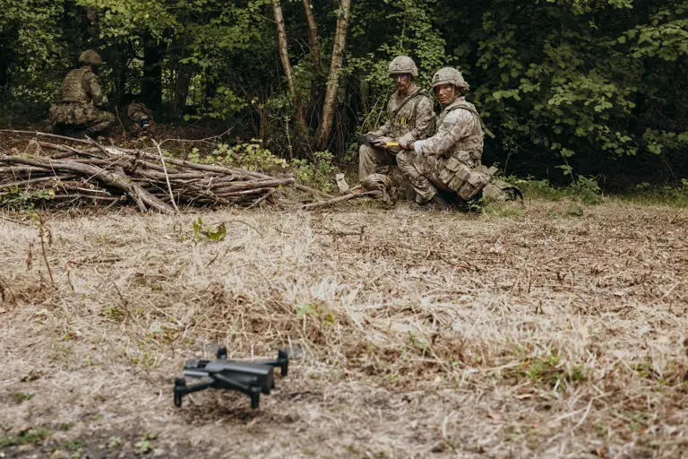 Two soldiers in camouflage gear kneel near the edge of a forest, operating a drone on dry grassland.