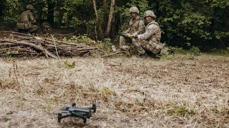 Two soldiers in camouflage gear kneel near the edge of a forest, operating a drone on dry grassland.