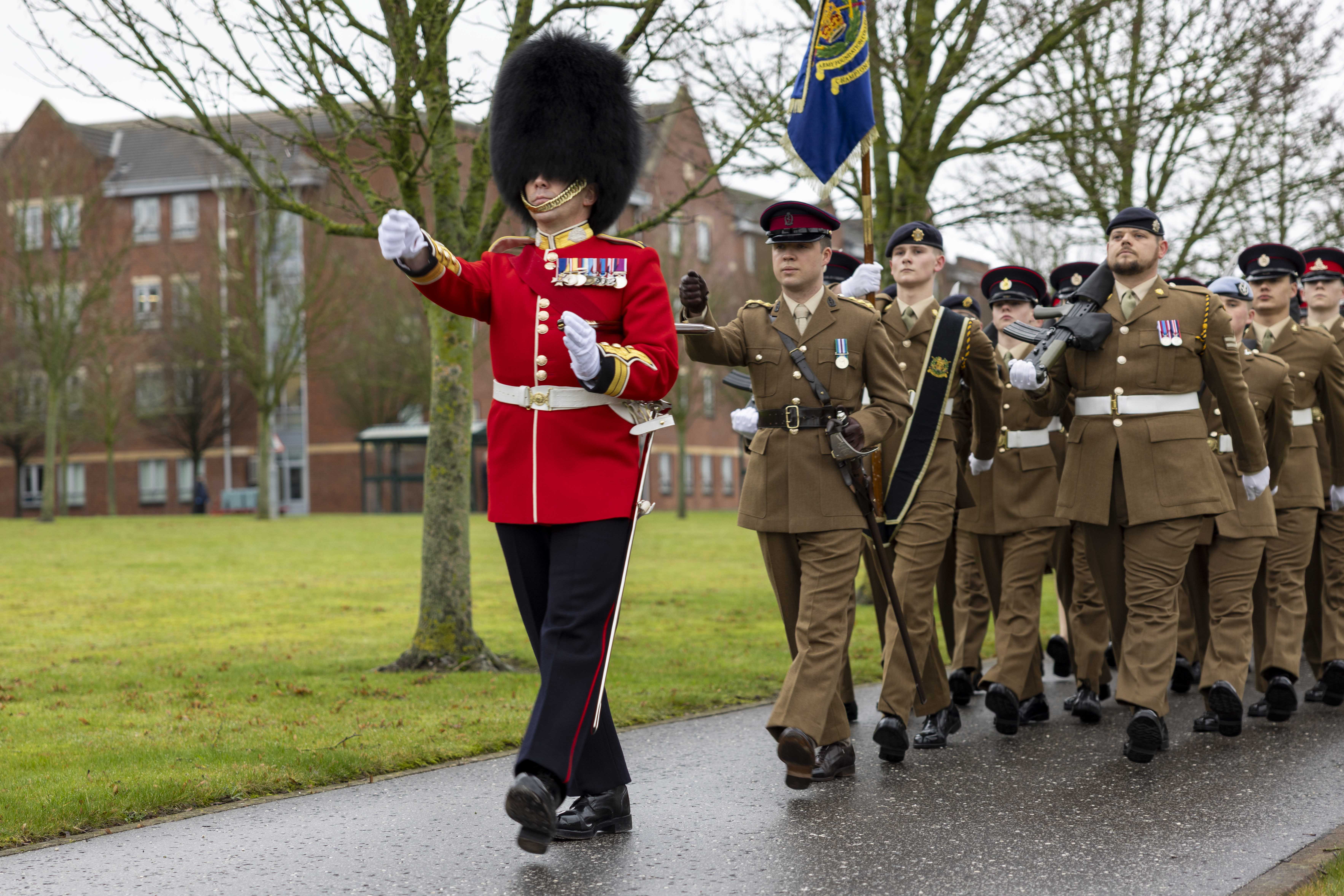 A military parade with a soldier in a red uniform and bearskin hat leads a formation of soldiers in brown uniforms holding a flag. Overcast day.