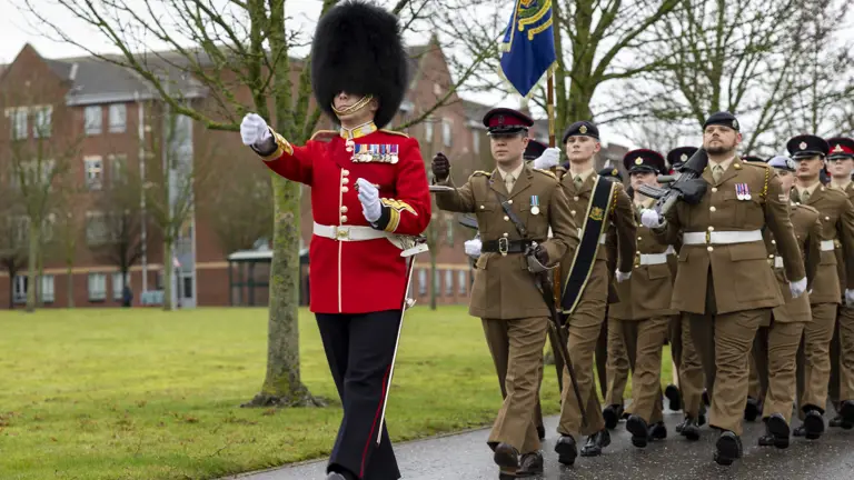 A military parade with a soldier in a red uniform and bearskin hat leads a formation of soldiers in brown uniforms holding a flag. Overcast day.