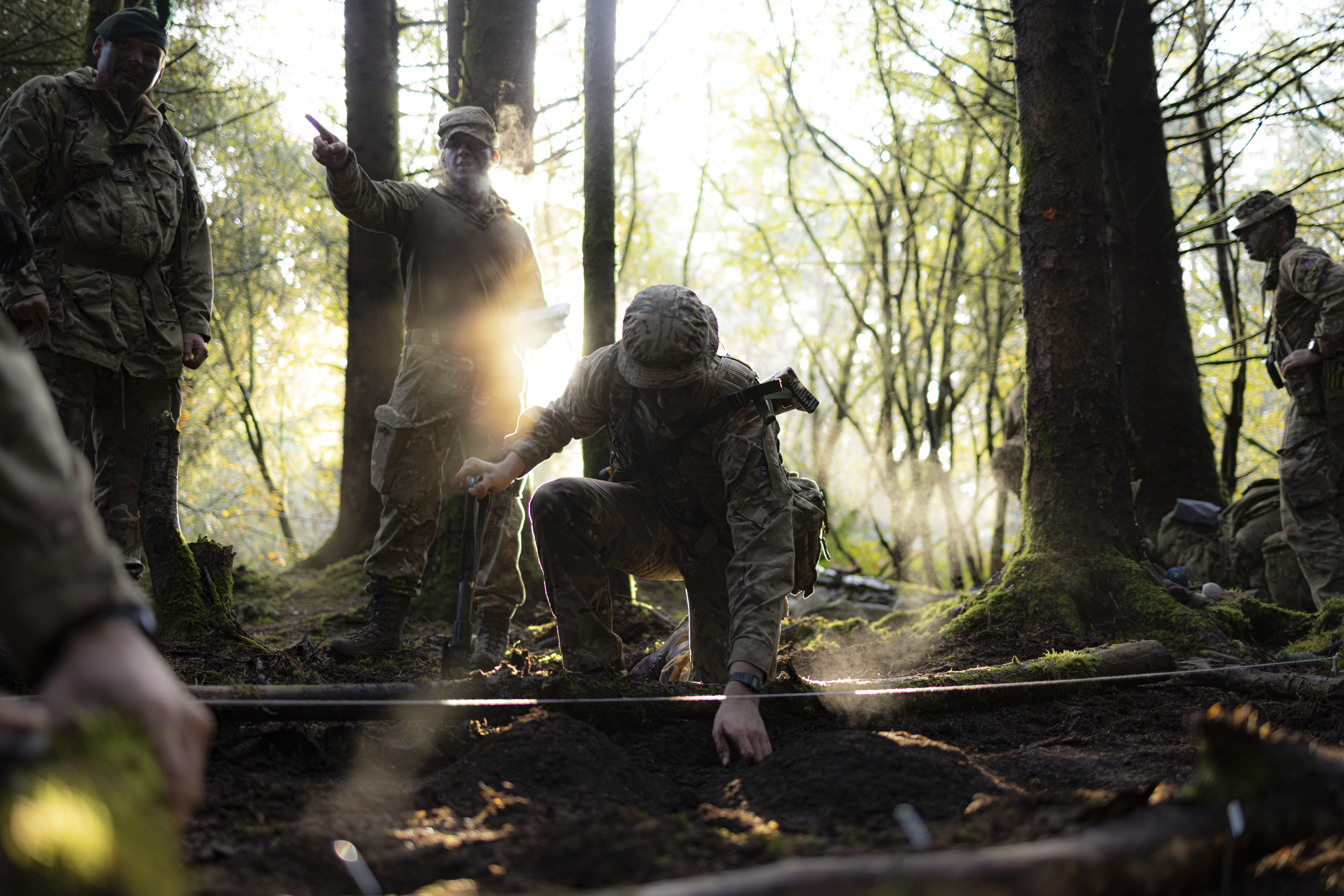 Several soldiers in uniform are pictured within a woodland on exercise. 