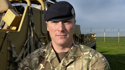 A British soldier, Sergeant Humphries, standing next to a military vehicle. He is wearing a camouflage uniform and in the background is a perimeter fence.