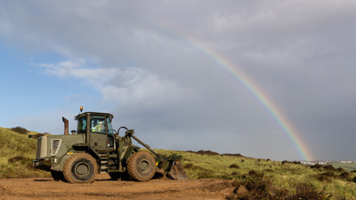 A green military digger is stripping away scrub on the top of a large sand dune. In the background a rainbow can be seen over the horizon.