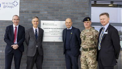 Five people are standing in front of a new building. Three of them are dressed in blue suits, one in a grey suite and the final is a soldier wearing camouflage and a beret. Two of them are standing to the left of a sign on the wall, marking the opening of the new building. The remaining three people are standing to the right of the sign.
