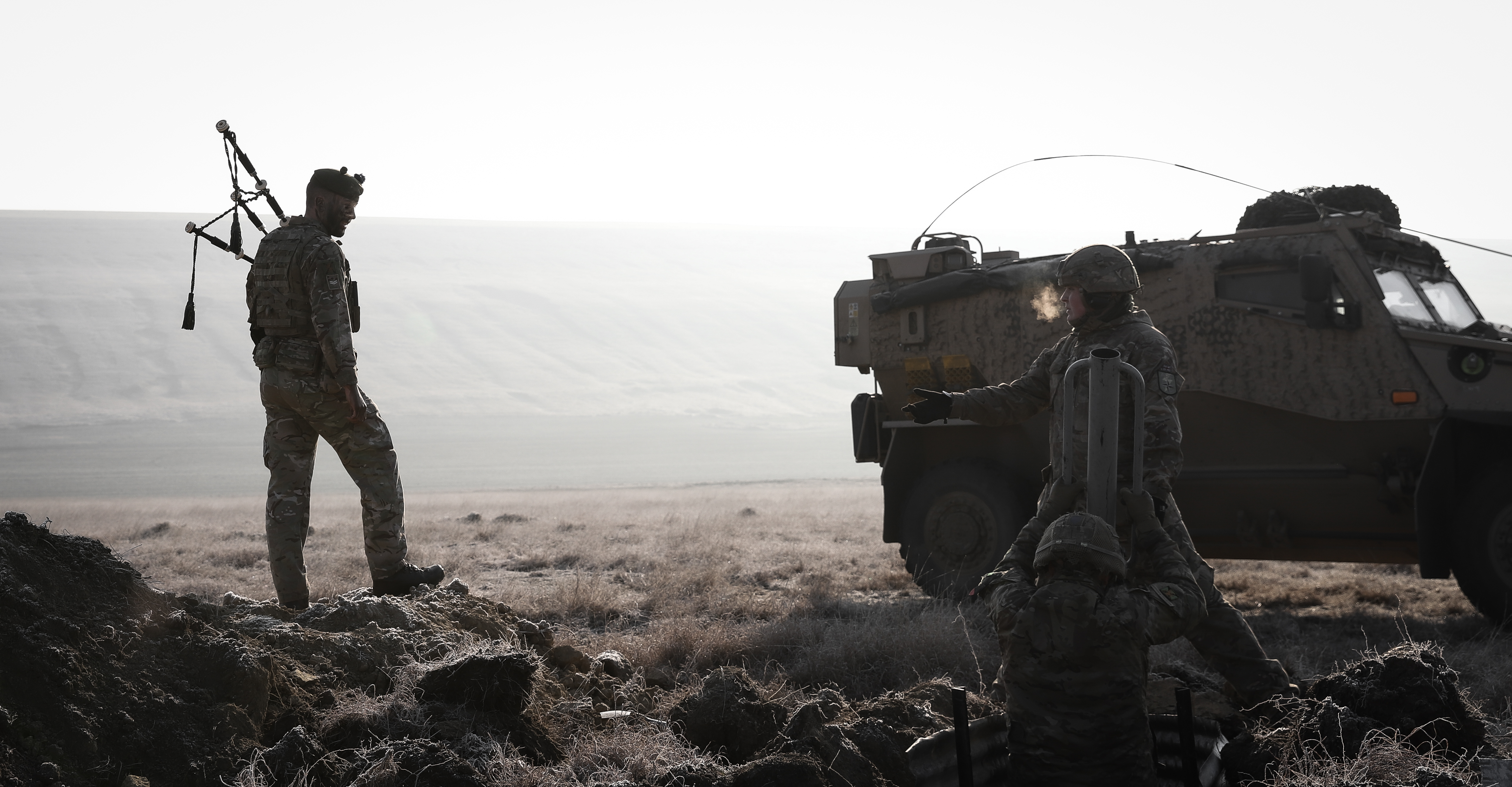 Image shows soldier in camouflage uniform in an active training field holding bagpipes while walking past other soldiers in body armour digging trenches in front of an army vehicle. 