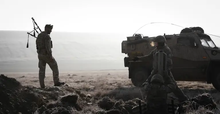 Image shows soldier in camouflage uniform in an active training field holding bagpipes while walking past other soldiers in body armour digging trenches in front of an army vehicle.
