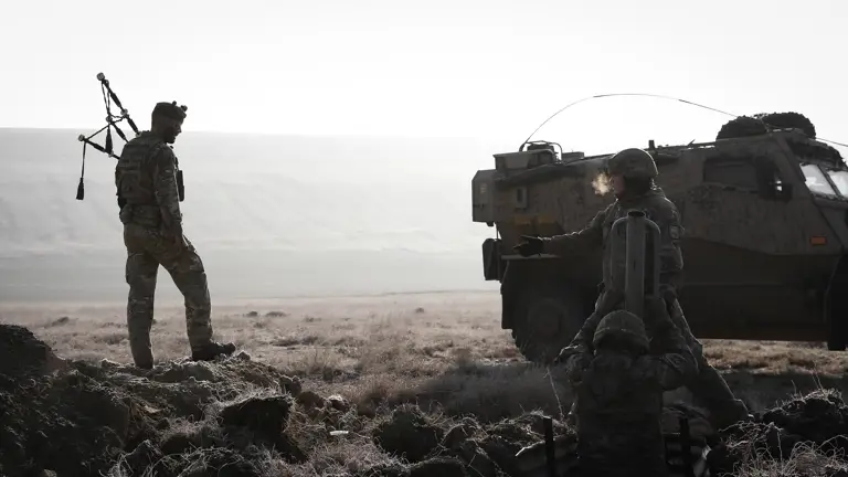 Image shows soldier in camouflage uniform in an active training field holding bagpipes while walking past other soldiers in body armour digging trenches in front of an army vehicle.