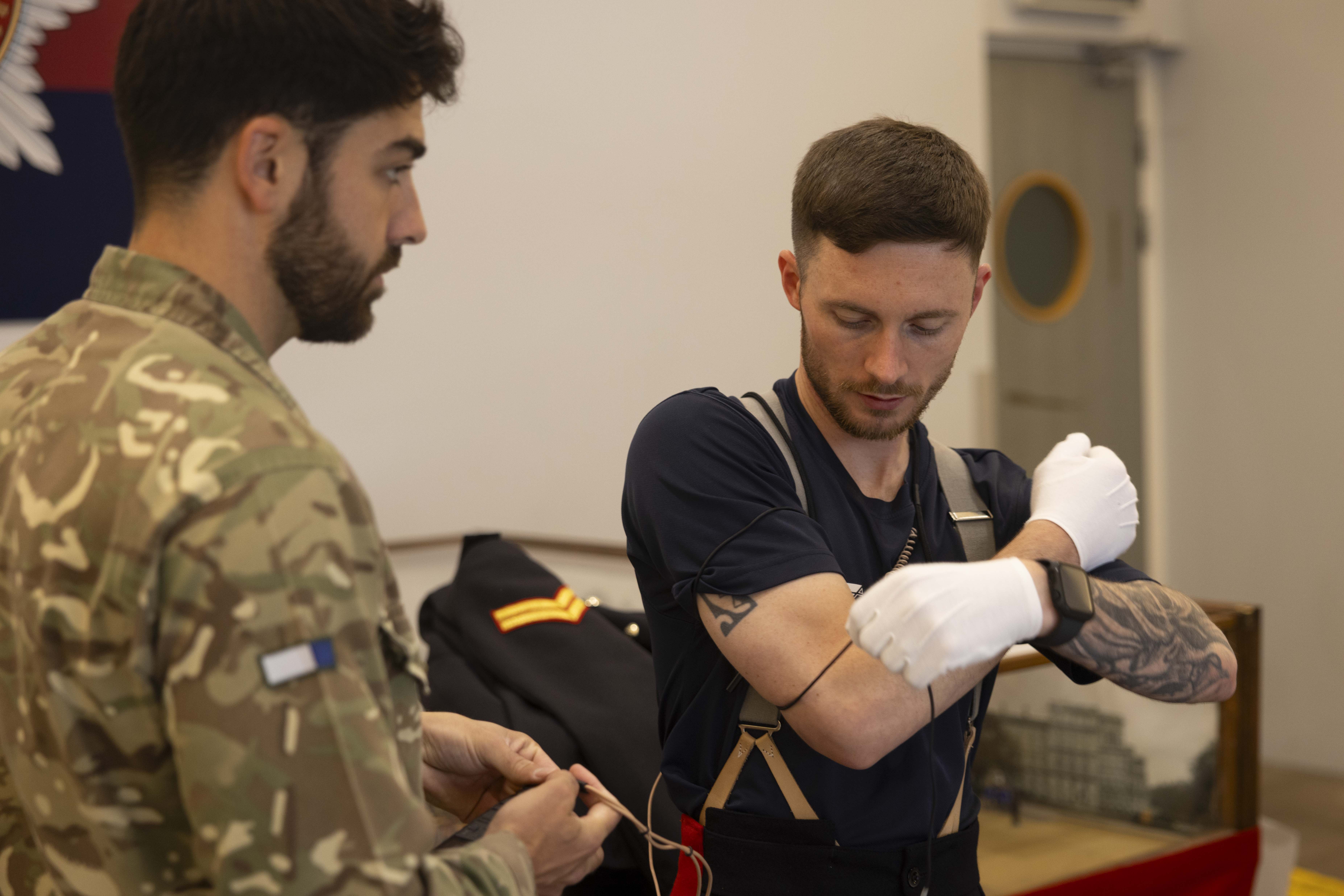 Two men are in a room. One wears a camouflage uniform, holding a wire, and the other, in casual attire, wearing gloves, appears focused on wiring his arm.