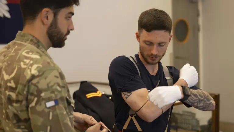 Two men are in a room. One wears a camouflage uniform, holding a wire, and the other, in casual attire, wearing gloves, appears focused on wiring his arm.