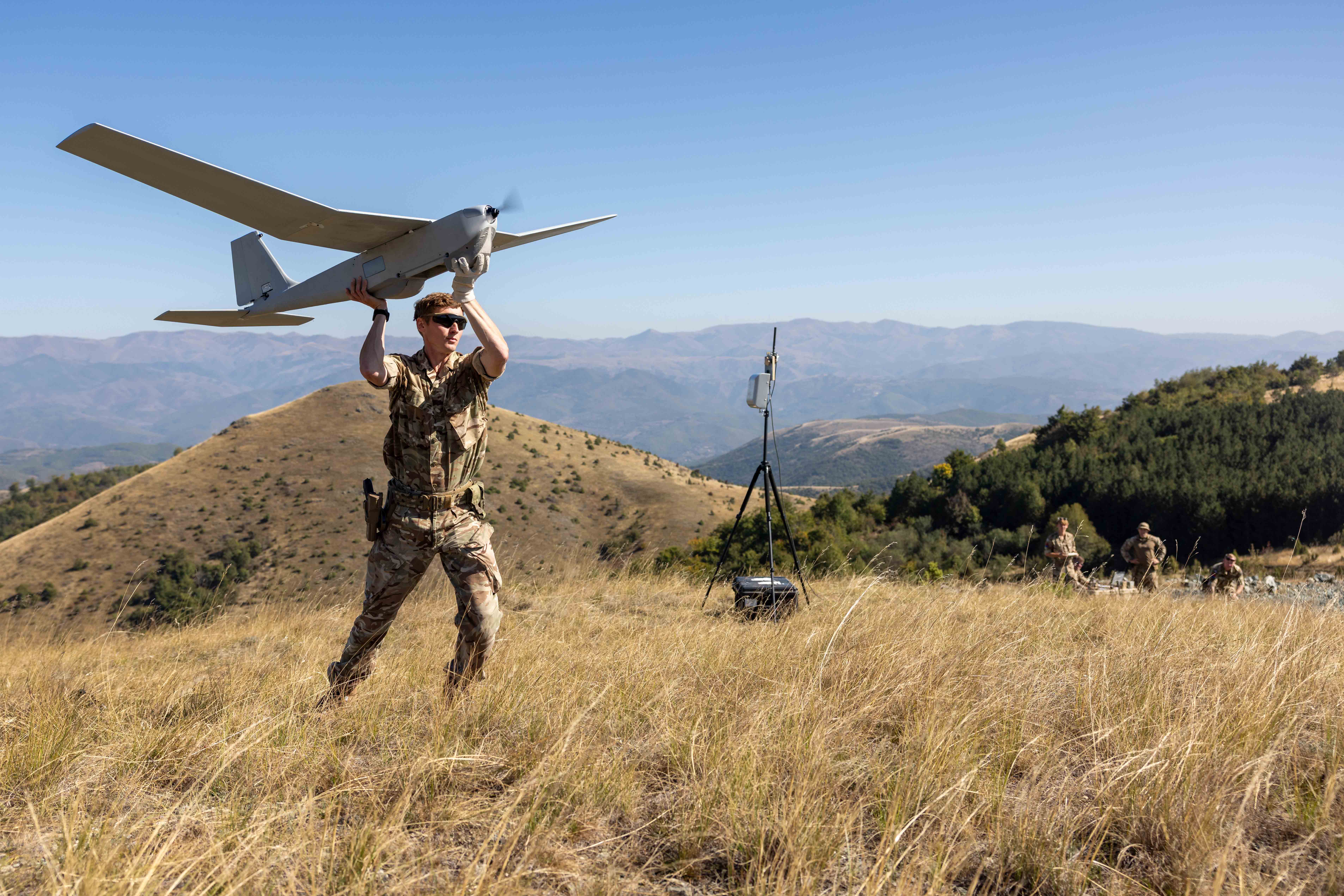 Lance Bombardier Morrissey is on the top of a hill wearing his camouflaged Army uniform and cap. He has both hands on an grey unmanned aerial system, which has a wing span of around 10 feet he is about to launch.