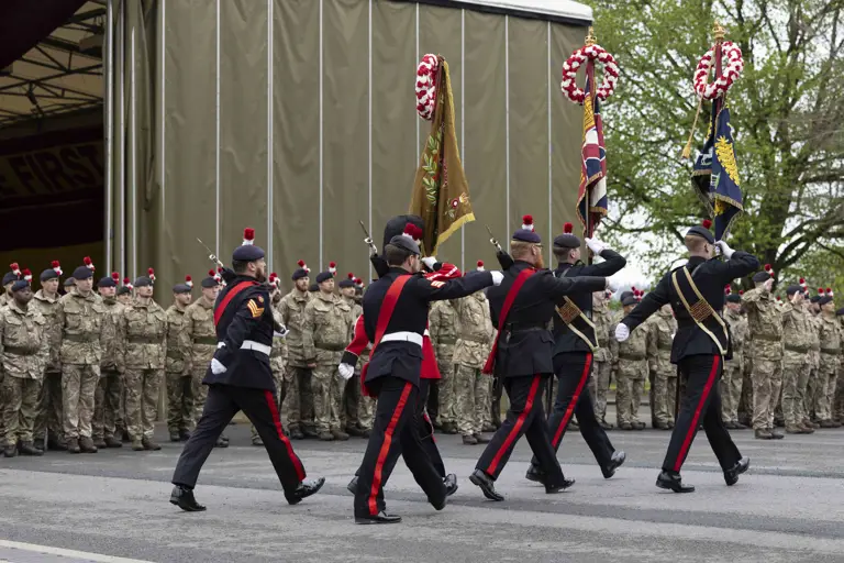 Soldiers in black ceremonial uniforms march past soldiers in green camouflage uniforms.