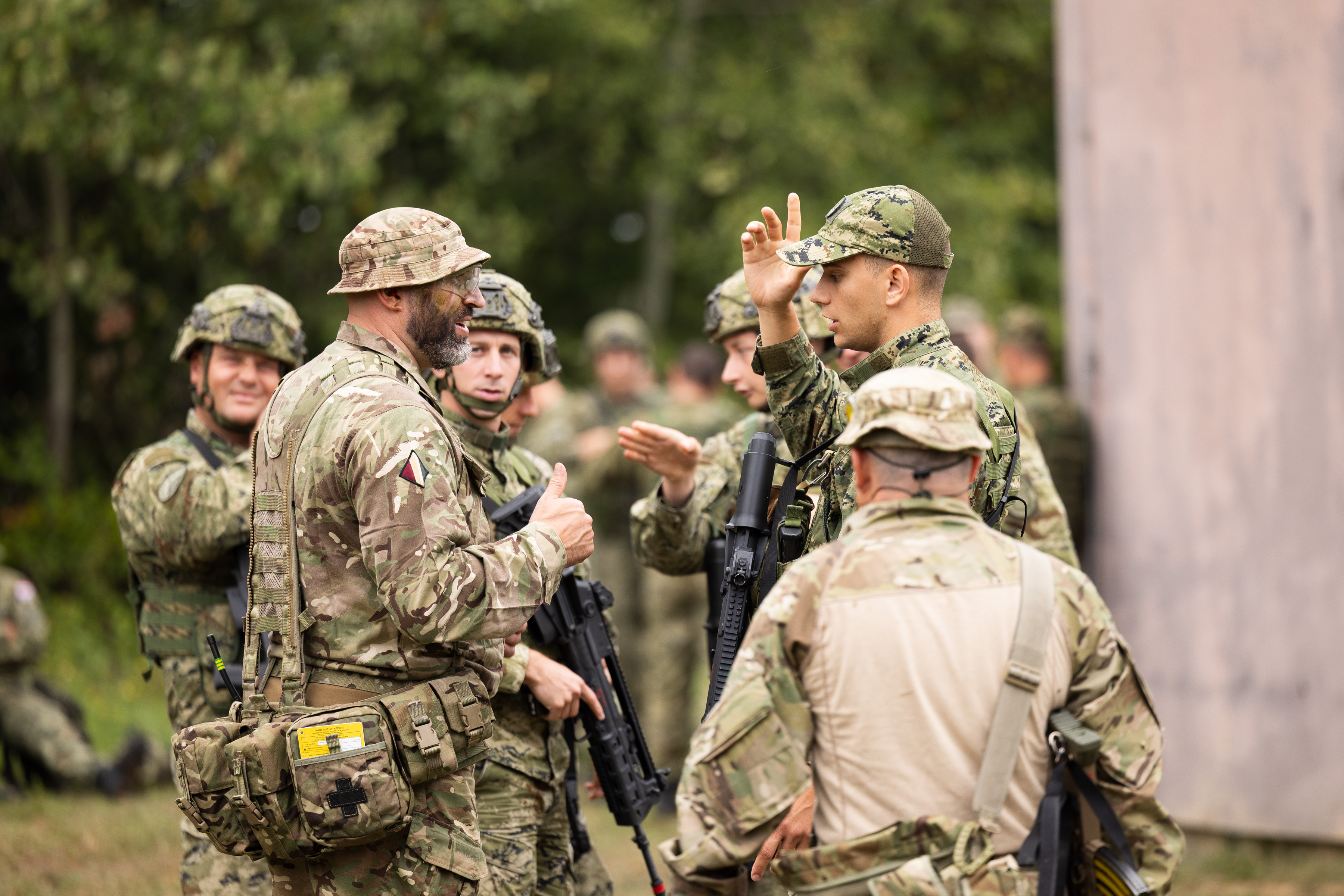 soldiers from both the British Army and the Croatian Armed Forces stand talking outside a building on exercise wearing their different camouflage uniforms 