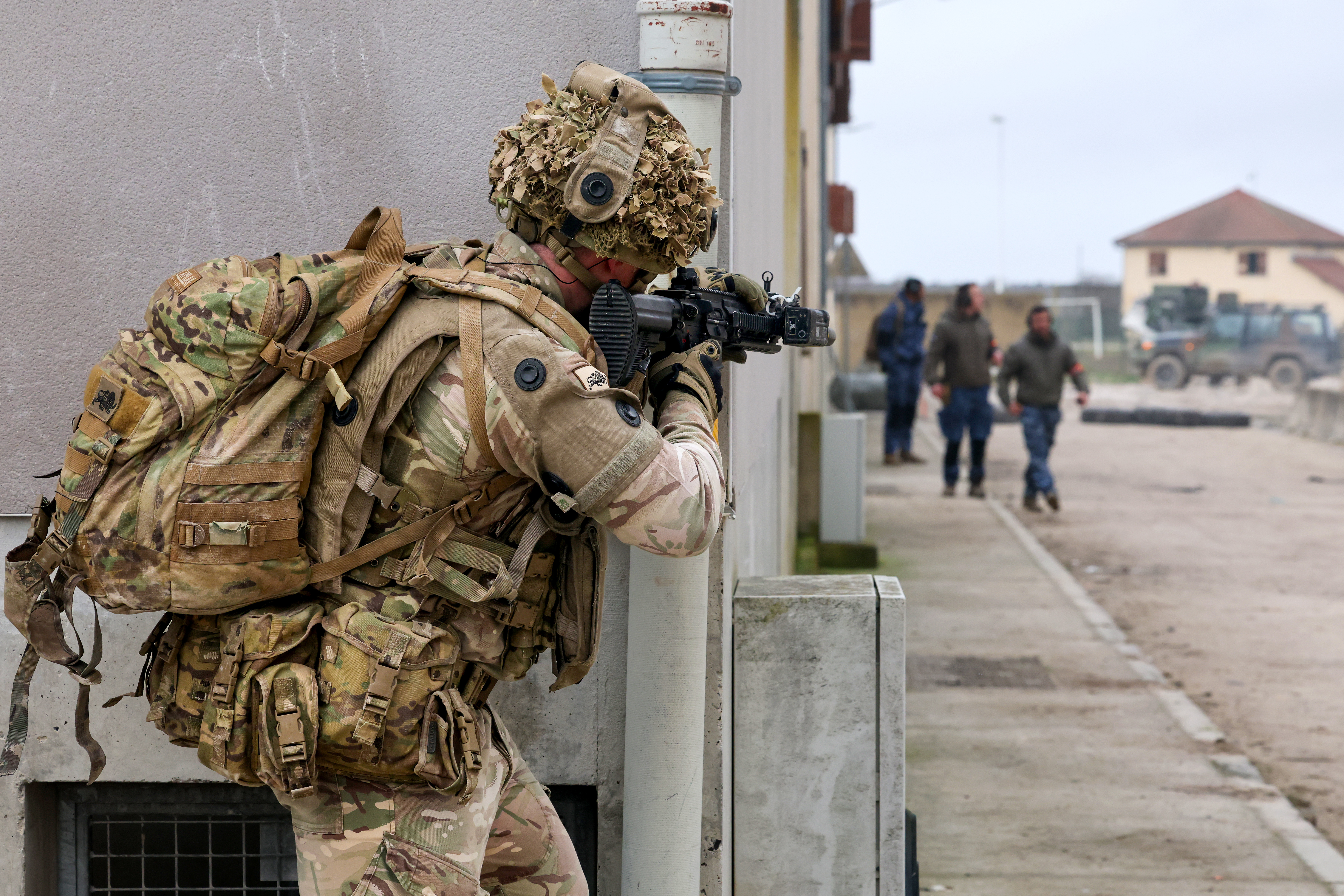 A person in military camouflage holds a rifle while taking cover behind a wall in an urban training area. 