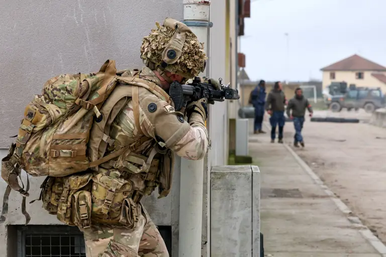 A person in military camouflage holds a rifle while taking cover behind a wall in an urban training area. 
