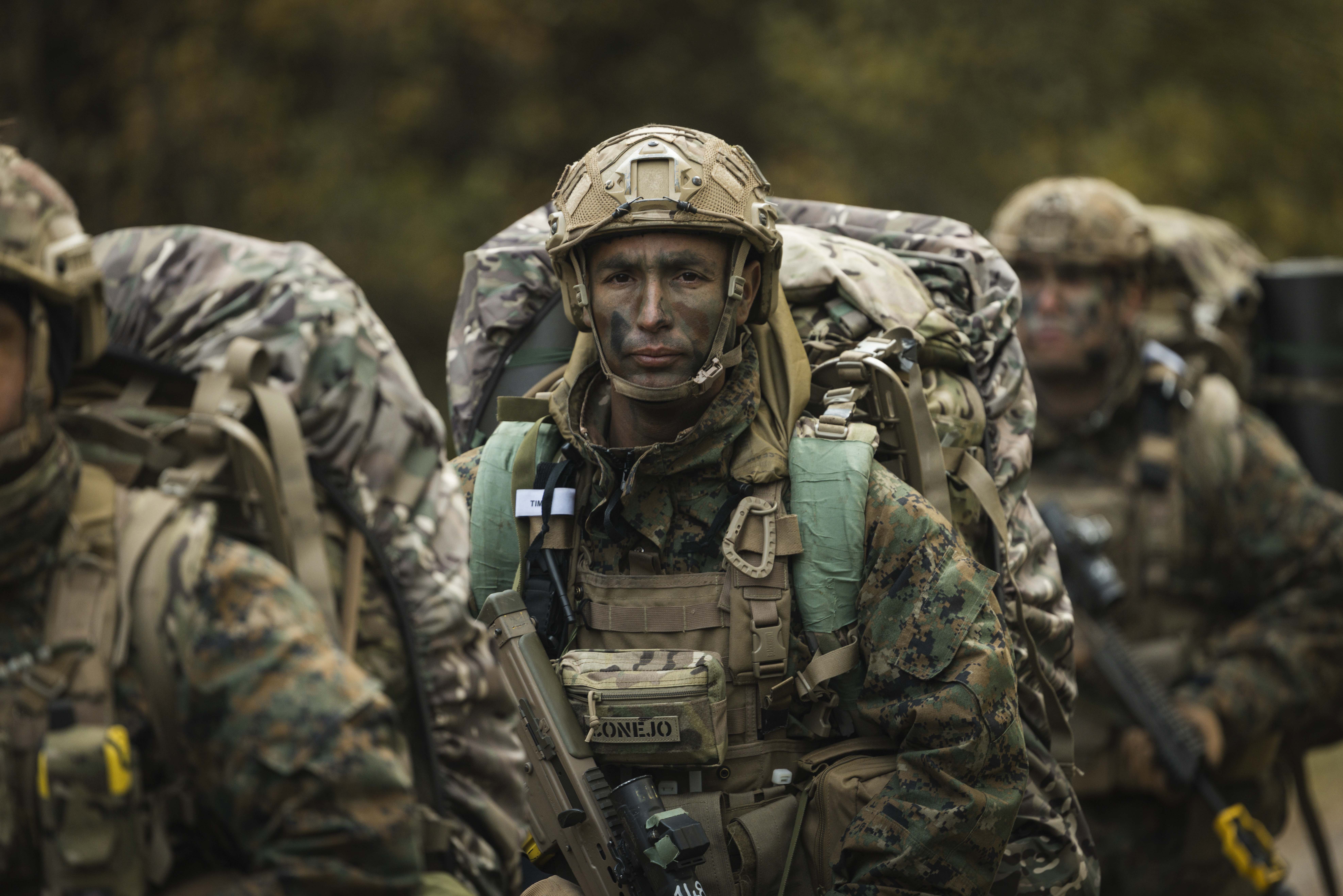 Soldiers in camouflage gear and helmets march through a forest, displaying determination. They carry large backpacks and rifles, conveying focus and readiness.